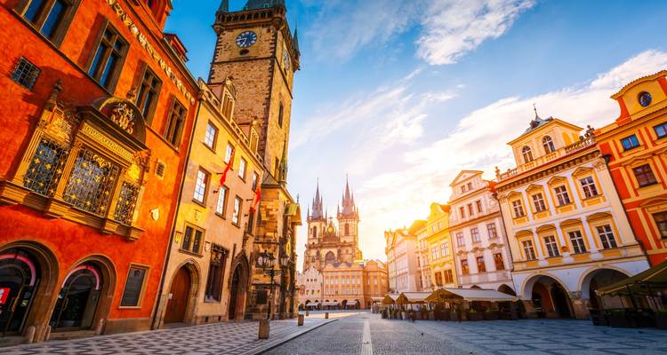 Cobblestoned square with ornate astronomical clock tower and spired church bathed in sunrise glow.