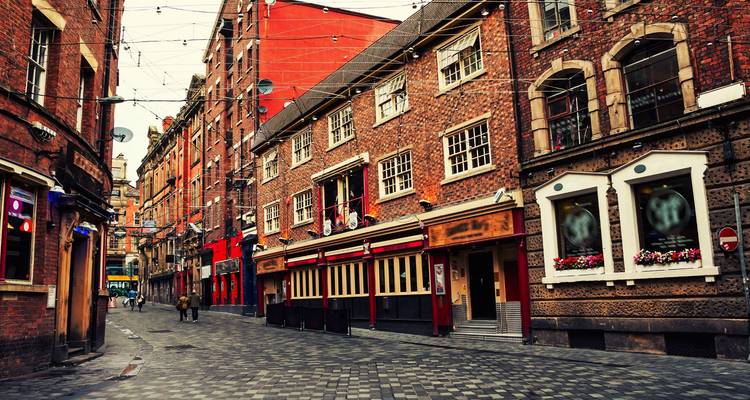 Narrow cobbled street lined with red-brick pubs and hanging lights in evening