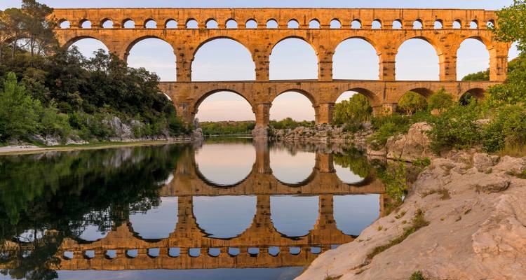 La lumière dorée du soir baigne les arches anciennes du Pont du Gard parfaitement reflétées dans la rivière en contrebas.