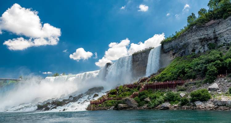 Vista amplia de las Cataratas Estadounidenses en el Niágara con niebla elevándose y senderos de madera abajo de un cielo azul brillante con nubes esponjosas.