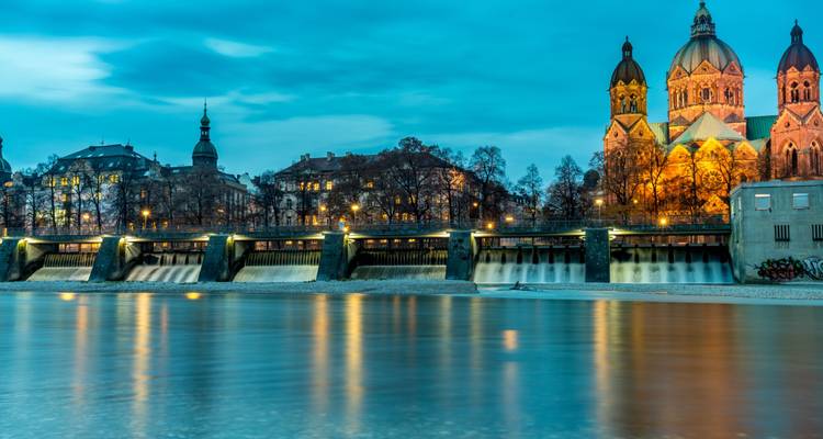 Blaue-Stunden-Blick auf die St.-Lukas-Kirche und das beleuchtete Wehr, das sich im ruhigen Isarfluss in München spiegelt