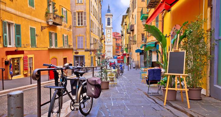 Rue piétonne colorée dans le Vieux Nice bordée de bâtiments aux couleurs pastel, de cafés, de vélos et d'aperçus d'une tour de l'horloge au loin.