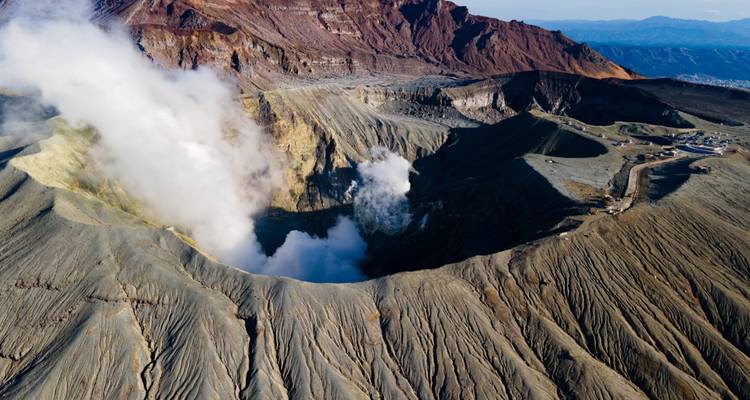 Vue aérienne d'un cratère volcanique fumant aux pentes striées accidentées et aux chaînes de montagnes environnantes.