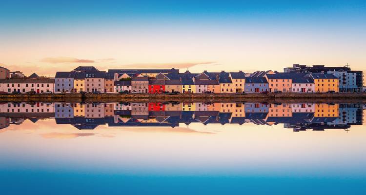 Reihe bunter Häuser, die sich perfekt in ruhigem Abendwasser unter pastellfarbenem Himmel in Galway spiegeln.