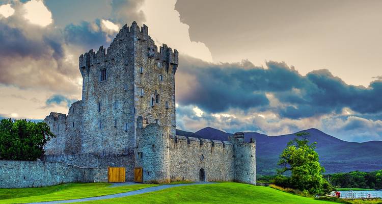 Stone Ross Castle ligt op een graspluik naast Lough Leane met zich uitstrekkende bergen en dramatische wolken erachter.