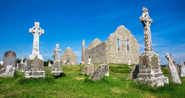 Des croix celtiques anciennes et des ruines en pierre du site monastique de Clonmacnoise sous un ciel bleu dégagé.