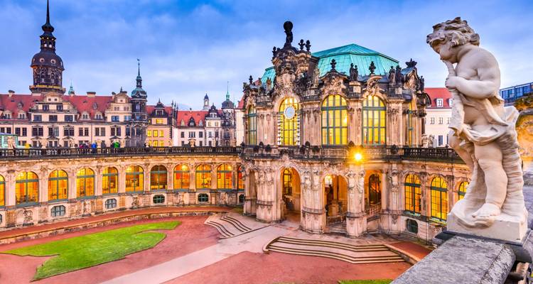 Der Barocke Dresdener Zwinger-Palast leuchtet in der Dämmerung mit warmem Innenlicht auf, betrachtet von seiner Balustrade mit einer Statue im Vordergrund.