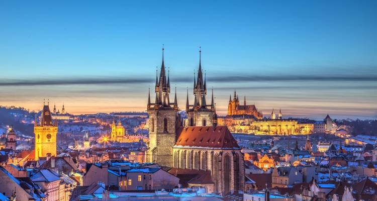 Dämmerung über Prags Altstadt mit schneebedeckten Dächern, die Zwillingstürme der Teynkirche und die beleuchtete Prager Burg in der Ferne.