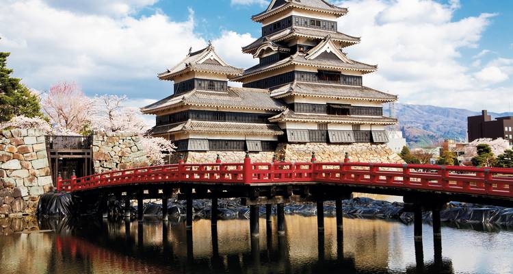 Le château de Matsumoto noir et blanc se reflétant dans ses douves derrière un pont en bois rouge et les fleurs du printemps.