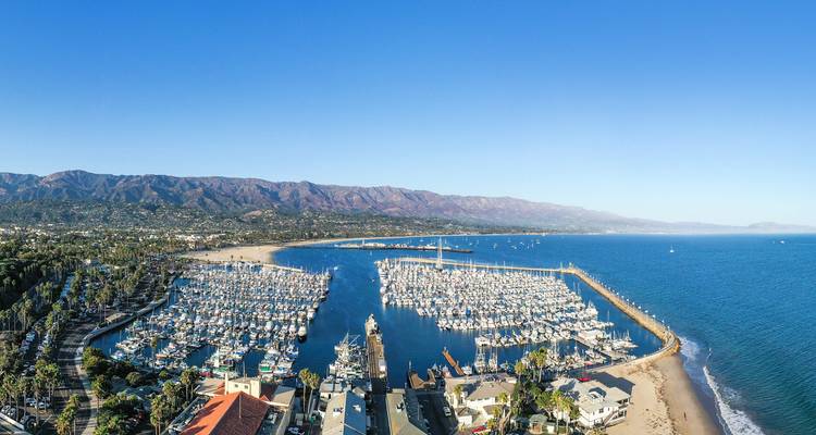 Vue panoramique aérienne d'une marina remplie de yachts s'incurvant dans le Pacifique à côté de sables dorés, avec des montagnes côtières s'étendant sous un ciel dégagé.