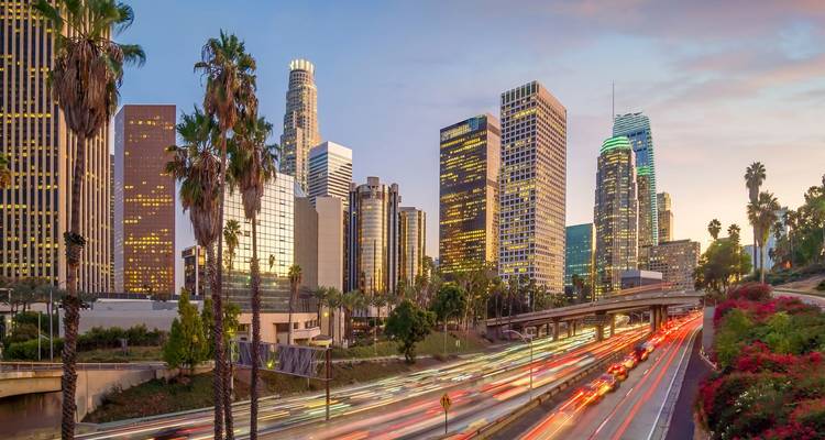 Les gratte-ciel du centre-ville de Los Angeles s'illuminent au crépuscule tandis que les phares des voitures qui circulent tracent des traînées lumineuses le long de l'autoroute bordée de palmiers.
