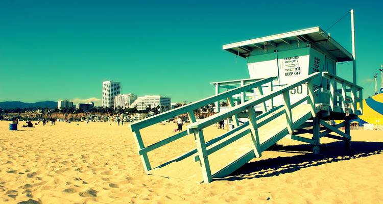 Une vue au ton rétro d'une cabine de maître-nageur bleu pâle surplombant une plage de sable animée avec des gratte-ciel urbains en arrière-plan.