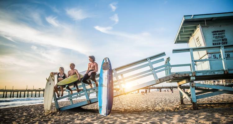 Trois jeunes surfeurs sont assis sur leurs planches à côté d'une tour de surveillance de maître-nageur au coucher du soleil, regardant vers les vagues douces et un horizon lumineux.