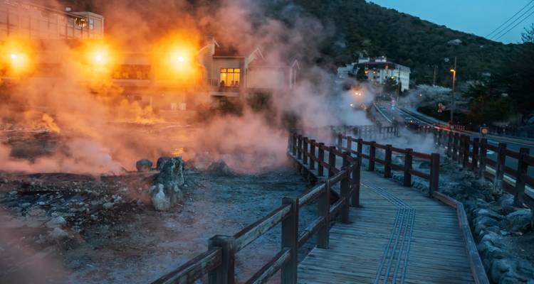 Steamy geothermal area at dusk with wooden boardwalk and glowing building lights.