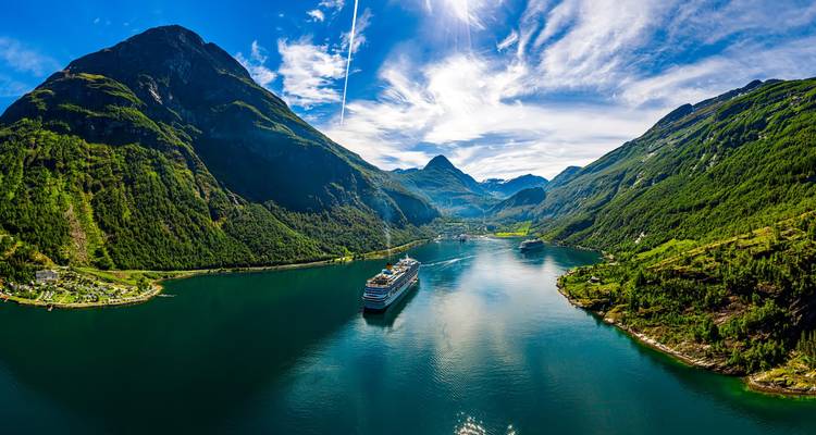 Vue aérienne du Geirangerfjord avec des montagnes verdoyantes imposantes et des navires de croisière glissant sur l'eau émeraude sous un ciel bleu éclatant.