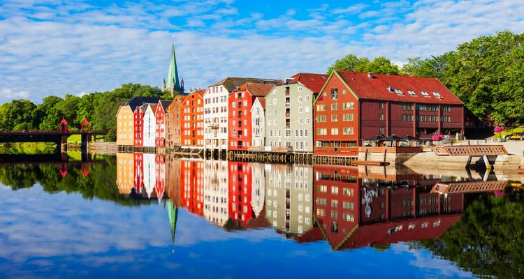 Les entrepôts aux couleurs vives bordant la rivière de Trondheim se reflétant vivement dans une eau calme et lisse sous un ciel parsemé.