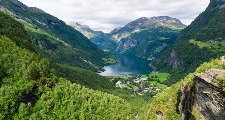Vue panoramique spectaculaire du Geirangerfjord aux eaux émeraude serpentant entre des montagnes escarpées et verdoyantes et un petit village en contrebas.
