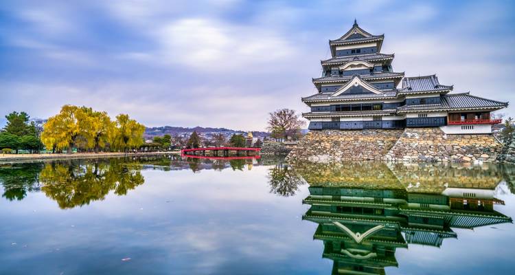 Vue reflétée du château de Matsumoto à côté d'un pont en arche rouge et d'arbres automnaux