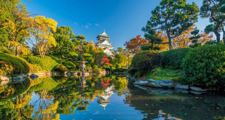 Bassin de jardin tranquille reflétant le château d'Osaka entouré d'arbres d'automne et de conifères sous un ciel bleu dégagé.