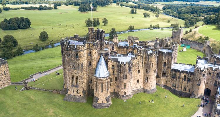 Perspective aérienne du château médiéval d'Alnwick entouré par la campagne verdoyante vallonnée et une rivière sinueuse.