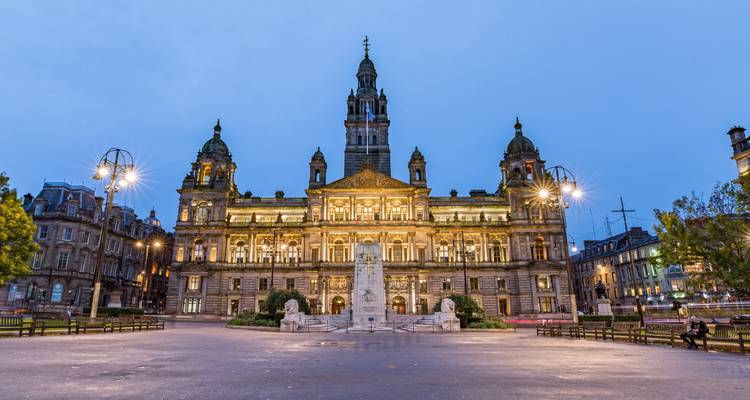 Les Chambres de la Ville de Glasgow illuminées dominent la Place George dans l'heure bleue avec des statues et des fontaines à l'avant.