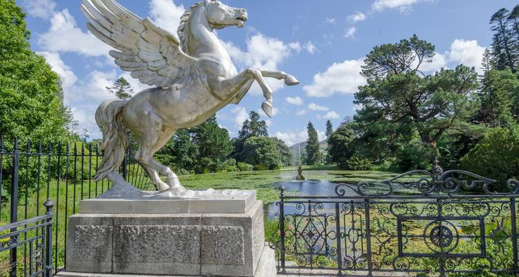 Statue de cheval ailé en marbre se cabrant sur un socle dominant un étang tranquille et des jardins entretenus sous un ciel bleu.