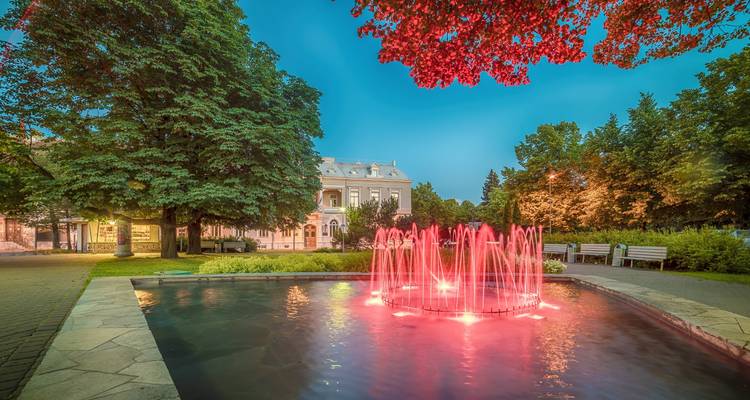 Escena nocturna de una pequeña piscina de parque con una fuente iluminada en rojo intenso, rodeada de árboles frondosos y una villa histórica.