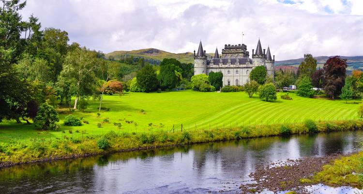 El Castillo de Inveraray está acurrucado en prados verdes perfectamente cuidados junto a un río que refleja la luz, con colinas boscosas detrás.