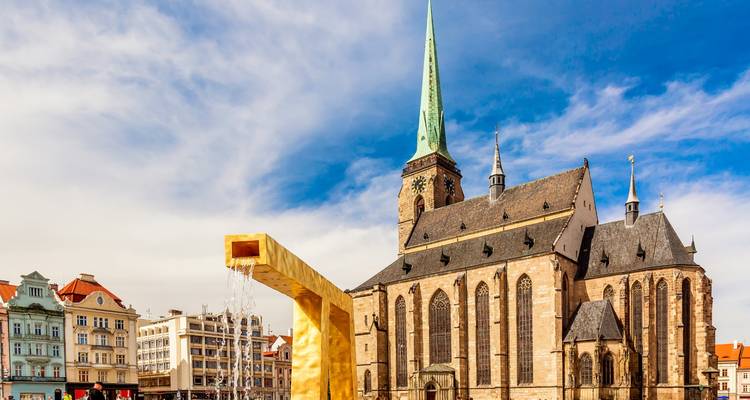 Place centrale de Pilsen avec la flèche de la cathédrale Sainte-Barthélemy et une sculpture de fontaine moderne dorée.
