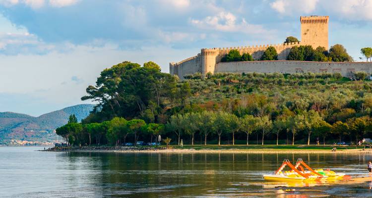 Forteresse médiévale au bord du lac sur une colline couverte d'arbres surplombant des eaux calmes et réfléchissantes avec des bateaux à pédales au premier plan.