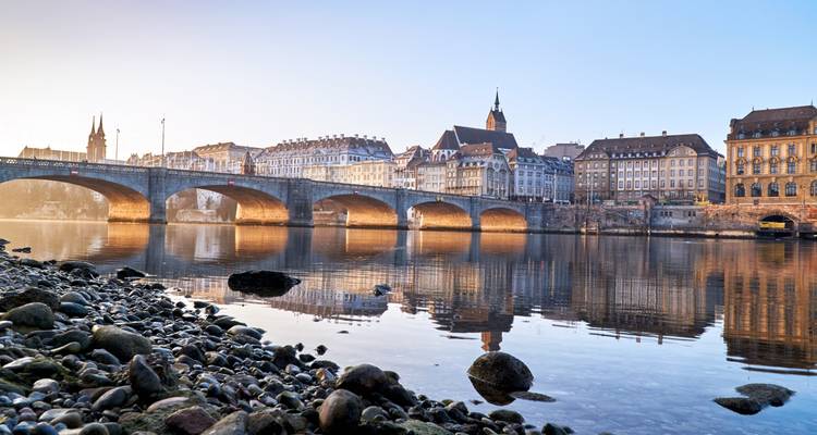 La luz del amanecer en el puente de piedra con arcos de Basilea y la orilla histórica del río reflejadas en el Rin.