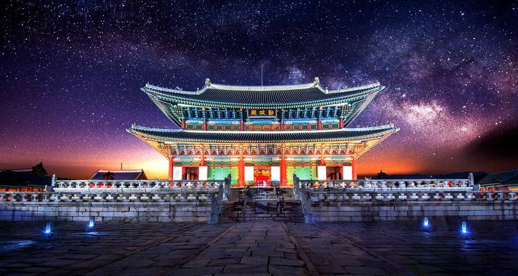 Vue nocturne d'un hall de palais coréen illuminé sous un ciel de Voie lactée rempli d'étoiles.