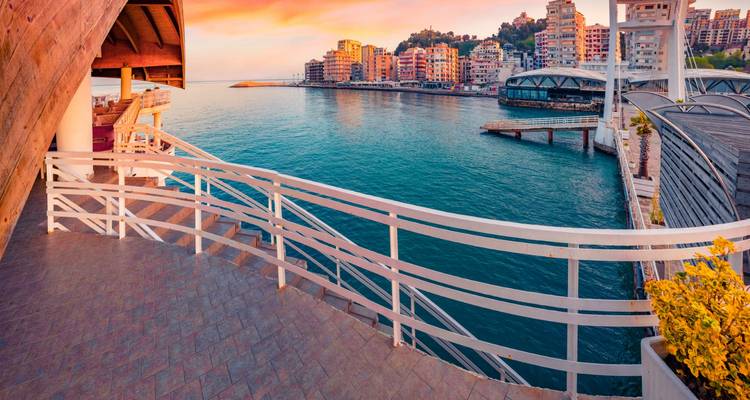 Vistas panorámicas de una terraza sobre una bahía turquesa salpicada de modernos bloques de apartamentos que suben por una ladera costera bajo un dramático cielo al atardecer.