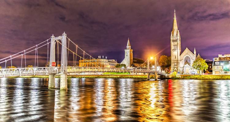 Vue nocturne du pont suspendu illuminé et des tours d'église se reflétant sur la rivière Ness