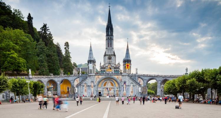 Esplanade, die zur imposanten Basilika Unserer Lieben Frau von Lourdes führt, umgeben von Pilgern und Besuchern unter einem düsteren Himmel.