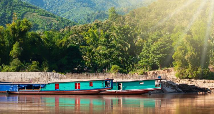 Lange hölzerne Frachtboote, die an einem ruhigen Mekong-Fluss vertäut sind, mit dschungelbedeckten Hügeln unter sanften Sonnenstrahlen.