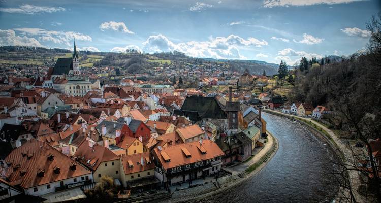 Historic town with red-tiled roofs winding along a curving river, surrounded by rolling hills under a partly cloudy sky.