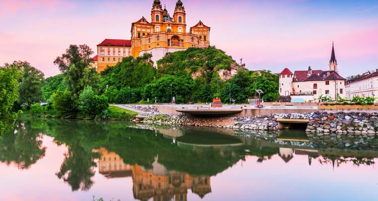 Abadía de Melk encaramada en una colina al atardecer con un cielo pastel, reflejada en el río tranquilo debajo.