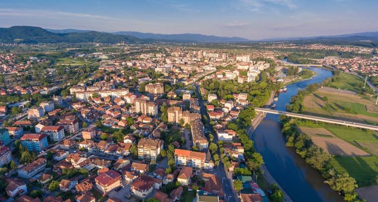Luftaufnahme einer mittelgroßen Stadt mit einem gewundenen Fluss, einer Brücke und grünen Hügeln unter klarem blauem Himmel.