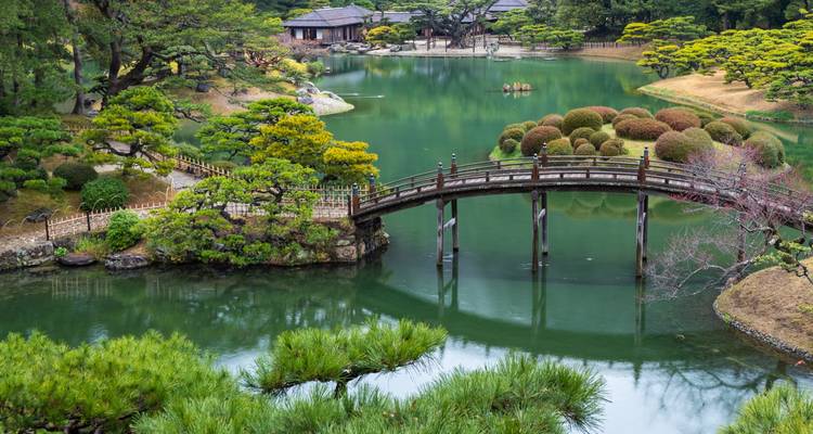 Serene Japanese landscape garden with an arched wooden bridge crossing emerald ponds, manicured pines and traditional teahouses.