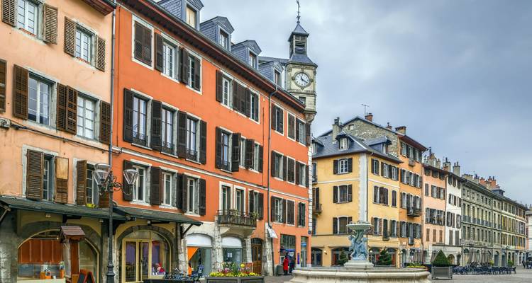 Une rangée colorée de bâtiments historiques et une fontaine centrale animent une rue piétonne dans la vieille ville de Chambéry.