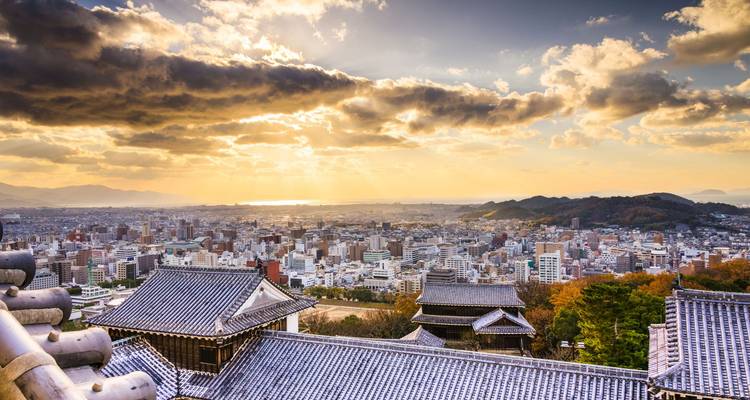 Vue panoramique du coucher de soleil sur les toits de la ville de Matsuyama depuis le château, avec des nuages dramatiques et la mer lointaine.