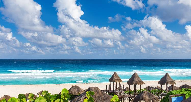 Escena de playa con palapas de paja, olas turquesas y una amplia orilla de arena.