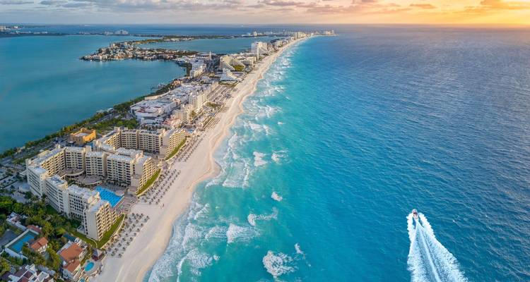 Panorama aéreo de la zona hotelera de Cancún con una larga playa blanca, mar turquesa y la estela de una lancha rápida.