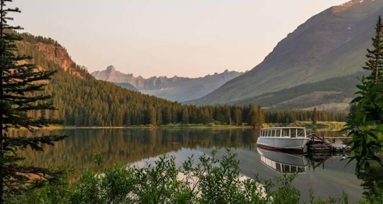 Lac de montagne pittoresque avec une forêt de conifères et un bateau de tourisme se reflétant sur l'eau calme sous une lumière chaude et matinale.