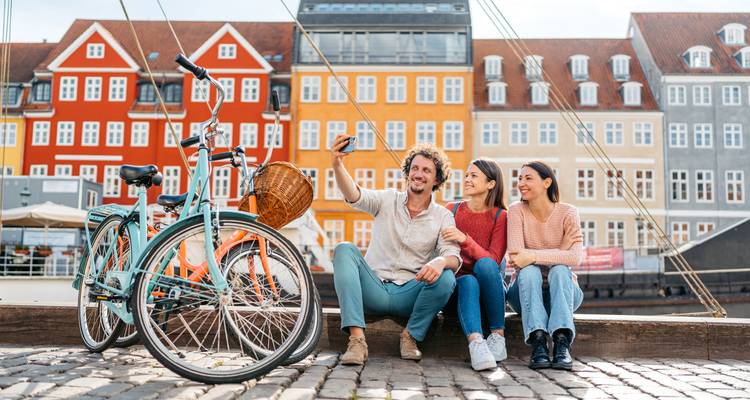 Trois amis sont assis sur un quai à Nyhavn avec des vélos tandis qu'ils se prennent en selfie devant des maisons danoises colorées.