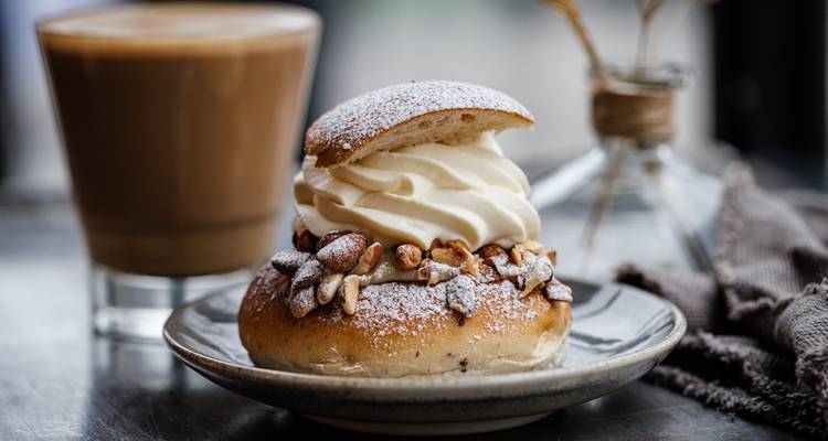 Nahaufnahme eines gepuderten Semla-Gebäcks gefüllt mit Sahne auf einem Teller neben einem Kaffeebecher.