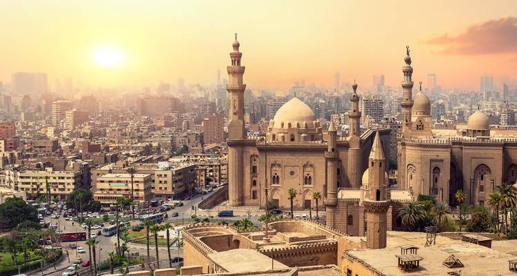 Goldener Sonnenaufgang über Kairos historischen Moscheen und Minaretten mit der dunstigen modernen Skyline dahinter.