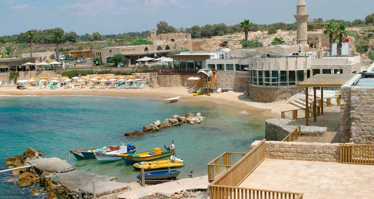 Sandige Bucht mit farbenfrohen Sonnenschirmen und Steingebäuden des antiken Hafens von Caesarea mit Minarett in Sichtweite.
