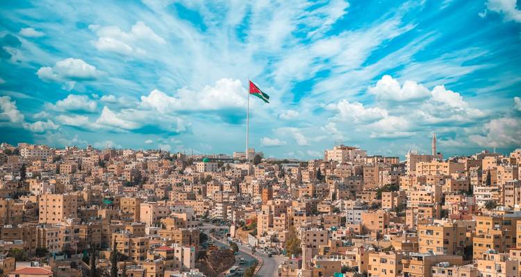 La ligne d'horizon d'Amman dominée par un immense mât du drapeau national sous des motifs nuageux bleu et blanc dramatiques.